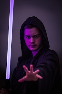 A person wearing a dark hooded robe holds a glowing purple lightsaber in their right hand, extending their left hand forward. The background is dark, emphasizing the mysterious and intense atmosphere.
