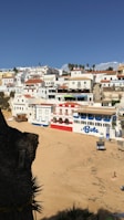 A picturesque coastal village with white buildings and colorful accents situated along a sandy beach. The architecture features red tile roofs, and the area appears serene under a clear blue sky. There is a prominent building labeled 'Bote' on the beachfront, and several palm trees in the distance add a touch of tropical ambiance.
