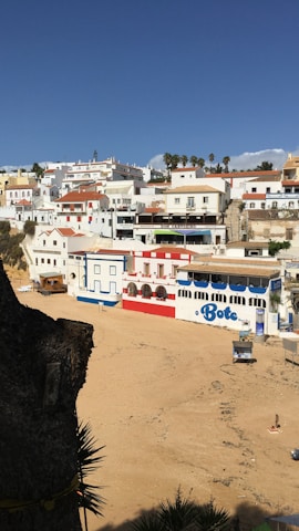 A picturesque coastal village with white buildings and colorful accents situated along a sandy beach. The architecture features red tile roofs, and the area appears serene under a clear blue sky. There is a prominent building labeled 'Bote' on the beachfront, and several palm trees in the distance add a touch of tropical ambiance.