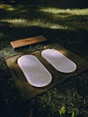 A set of white acupressure mats with spikes rests on a green cloth, placed on a grassy field. Adjacent to them, there is a rectangular wooden block with a design engraved on it.