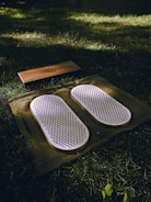A set of white acupressure mats with spikes rests on a green cloth, placed on a grassy field. Adjacent to them, there is a rectangular wooden block with a design engraved on it.