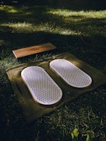 A set of white acupressure mats with spikes rests on a green cloth, placed on a grassy field. Adjacent to them, there is a rectangular wooden block with a design engraved on it.