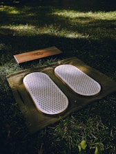 A set of white acupressure mats with spikes rests on a green cloth, placed on a grassy field. Adjacent to them, there is a rectangular wooden block with a design engraved on it.