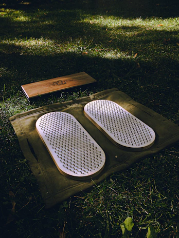 A set of white acupressure mats with spikes rests on a green cloth, placed on a grassy field. Adjacent to them, there is a rectangular wooden block with a design engraved on it.