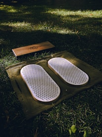 A set of white acupressure mats with spikes rests on a green cloth, placed on a grassy field. Adjacent to them, there is a rectangular wooden block with a design engraved on it.