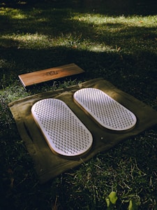 A set of white acupressure mats with spikes rests on a green cloth, placed on a grassy field. Adjacent to them, there is a rectangular wooden block with a design engraved on it.