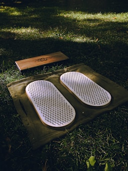 A set of white acupressure mats with spikes rests on a green cloth, placed on a grassy field. Adjacent to them, there is a rectangular wooden block with a design engraved on it.
