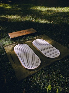 A set of white acupressure mats with spikes rests on a green cloth, placed on a grassy field. Adjacent to them, there is a rectangular wooden block with a design engraved on it.