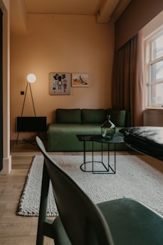 A cozy living room with warm lighting. The room features a green sofa against a light-colored wall, decorated with two framed artworks. A modern floor lamp with a spherical bulb stands to the side, providing ambient light. A small black metal frame table with a glass bottle sits on a textured area rug, and natural light streams in through a window with heavy drapes. A wooden floor and a modern-style chair add to the minimalist aesthetic.