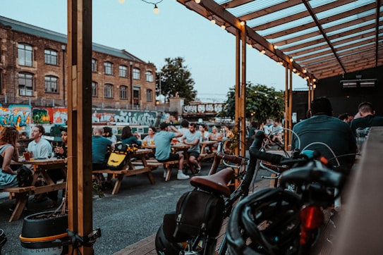 An outdoor urban setting featuring several groups of people dining at wooden picnic benches under a wooden canopy with string lights. Bicycles are parked in the foreground, and graffiti art can be seen on brick walls in the background, adding a lively and artistic vibe to the atmosphere.