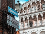 A detailed view of an ornate historical building with intricate stone carvings and arches. Street signs pointing to Palazzo Pfanner and Museo Casa Puccini are visible, set against a backdrop of brick and stone architecture. A vintage-style street lamp hangs near the signs.