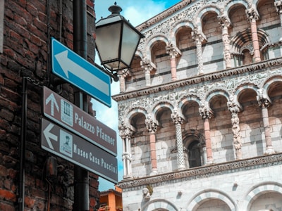 A detailed view of an ornate historical building with intricate stone carvings and arches. Street signs pointing to Palazzo Pfanner and Museo Casa Puccini are visible, set against a backdrop of brick and stone architecture. A vintage-style street lamp hangs near the signs.