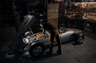 A person works intently on a vintage car with its hood open in a dimly lit garage. Tools and various mechanical parts are organized on shelves and walls, casting a warm, focused atmosphere. The vehicle appears to be an older model with a silver body and exposed engine components.
