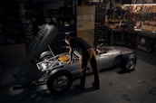 A person works intently on a vintage car with its hood open in a dimly lit garage. Tools and various mechanical parts are organized on shelves and walls, casting a warm, focused atmosphere. The vehicle appears to be an older model with a silver body and exposed engine components.
