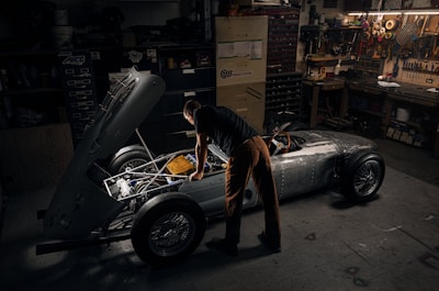 A person works intently on a vintage car with its hood open in a dimly lit garage. Tools and various mechanical parts are organized on shelves and walls, casting a warm, focused atmosphere. The vehicle appears to be an older model with a silver body and exposed engine components.
