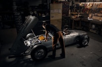 A person works intently on a vintage car with its hood open in a dimly lit garage. Tools and various mechanical parts are organized on shelves and walls, casting a warm, focused atmosphere. The vehicle appears to be an older model with a silver body and exposed engine components.