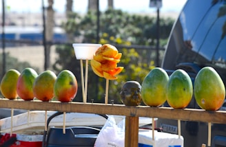 A row of ripe mangoes is displayed on a wooden stand. In the middle, a cup holds a peeled and sliced mango seasoned with a red spice. Next to the cup is a small statue resembling a human head. The background reveals a blurry outdoor setting with greenery and vehicles.