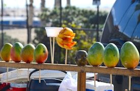A row of ripe mangoes is displayed on a wooden stand. In the middle, a cup holds a peeled and sliced mango seasoned with a red spice. Next to the cup is a small statue resembling a human head. The background reveals a blurry outdoor setting with greenery and vehicles.