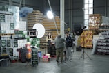 A warehouse setting with stacks of product boxes forming a backdrop. Several lighting and camera equipment pieces are set up, suggesting a photo or video shoot. Three people are present, two adjusting equipment and one sitting at a table with various items on it. The area is well-organized, and the lighting is bright.