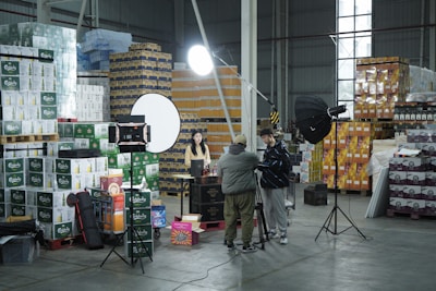 A warehouse setting with stacks of product boxes forming a backdrop. Several lighting and camera equipment pieces are set up, suggesting a photo or video shoot. Three people are present, two adjusting equipment and one sitting at a table with various items on it. The area is well-organized, and the lighting is bright.