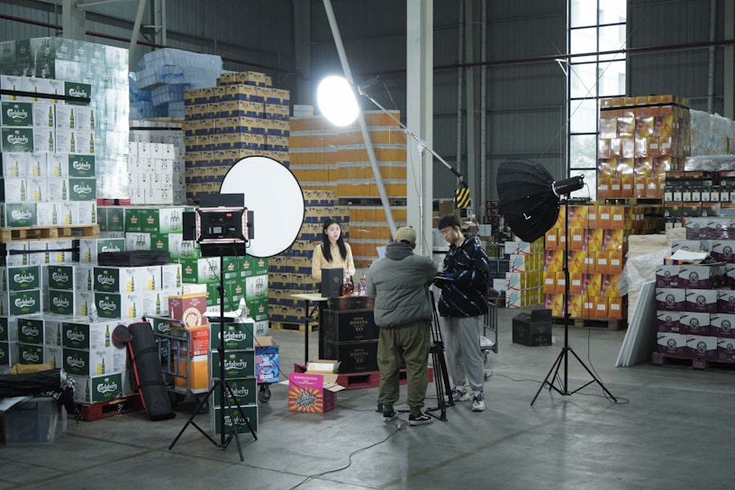 A warehouse setting with stacks of product boxes forming a backdrop. Several lighting and camera equipment pieces are set up, suggesting a photo or video shoot. Three people are present, two adjusting equipment and one sitting at a table with various items on it. The area is well-organized, and the lighting is bright.