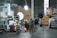 A warehouse setting with stacks of product boxes forming a backdrop. Several lighting and camera equipment pieces are set up, suggesting a photo or video shoot. Three people are present, two adjusting equipment and one sitting at a table with various items on it. The area is well-organized, and the lighting is bright.