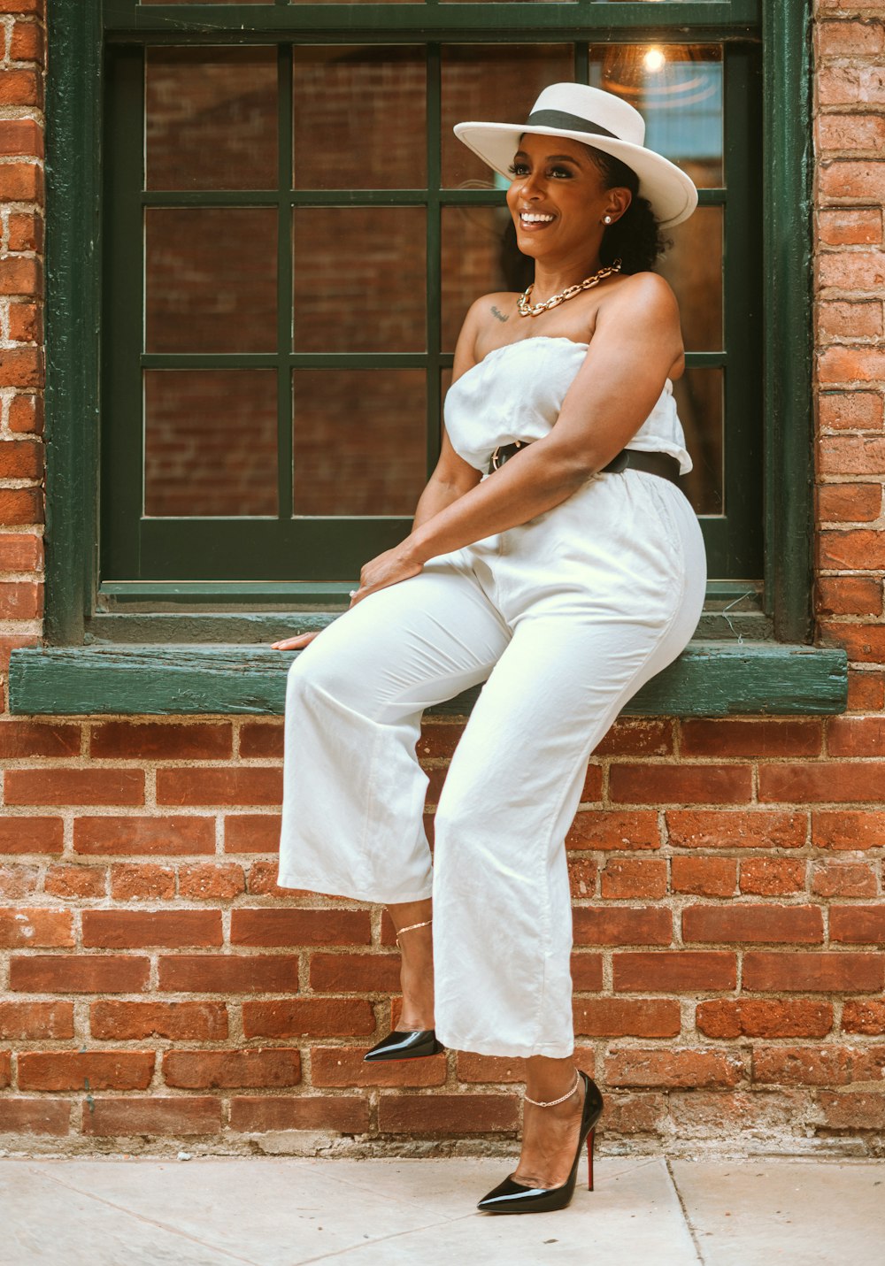 woman in white tank top and white pants leaning on brown brick wall