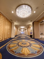 A luxurious hallway with ornate chandeliers hanging from the ceiling. The walls are decorated with intricate patterns, and the floor is covered with an elaborately designed carpet in rich blue, gold, and brown tones. Recessed lighting adds to the opulence of the setting.