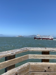 A large ferry boat sails across a body of water under a clear blue sky. In the foreground, a wooden railing juts out over the water, showing weathered planks with visible bolts and slight graffiti. In the distance, an island and mountainous shoreline are visible.