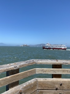 A large ferry boat sails across a body of water under a clear blue sky. In the foreground, a wooden railing juts out over the water, showing weathered planks with visible bolts and slight graffiti. In the distance, an island and mountainous shoreline are visible.
