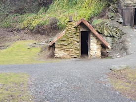 A small, rustic stone cottage with a slanted red-tiled roof is nestled into a hillside. The entrance is dark and open, with a rough stone path leading up to it. The surrounding area is covered with a mix of patches of green grass and bare soil, with some vegetation growing on the hillside.