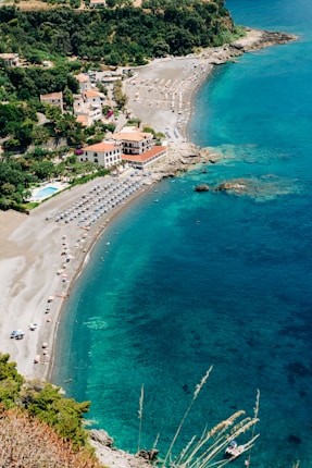 A scenic coastal area with crystal-clear turquoise waters and a narrow, curved beach lined with sun loungers and colorful umbrellas. A cluster of buildings with terra-cotta roofs is situated next to the beach, surrounded by lush greenery. The area is serene, with a few small boats visible in the water.