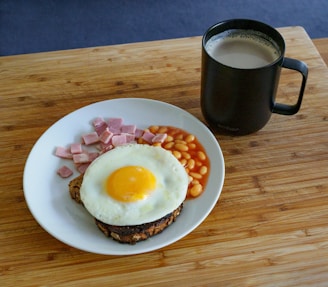 Plate containing a breakfast meal with a fried egg on top of toast, accompanied by diced ham and baked beans. A black mug filled with a hot beverage is placed on a wooden surface next to the plate.