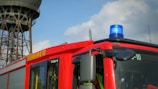 A bright red fire truck with a blue emergency light on top. The side of the truck is marked with 'FEUERWEHR 112' in bold yellow letters. A large, industrial-looking tower is in the background against a partly cloudy sky.