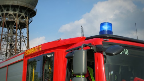A bright red fire truck with a blue emergency light on top. The side of the truck is marked with 'FEUERWEHR 112' in bold yellow letters. A large, industrial-looking tower is in the background against a partly cloudy sky.
