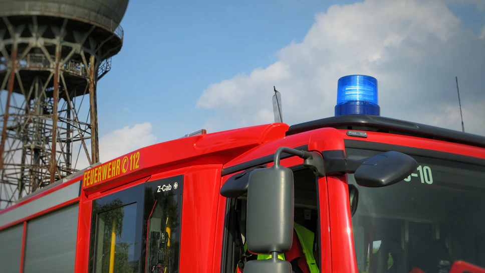 A bright red fire truck with a blue emergency light on top. The side of the truck is marked with 'FEUERWEHR 112' in bold yellow letters. A large, industrial-looking tower is in the background against a partly cloudy sky.