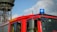 A bright red fire truck with a blue emergency light on top. The side of the truck is marked with 'FEUERWEHR 112' in bold yellow letters. A large, industrial-looking tower is in the background against a partly cloudy sky.