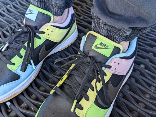A pair of colorful Nike sneakers on a textured rubber mat. The shoes combine pastel colors, including light blue, green, and pink, with black accents. The Nike logo is prominently displayed on the tongue, and the shoes are laced with black laces.