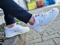 A pair of white sneakers with colorful paint splatters, worn by a person sitting on a low concrete ledge. The sneakers have a classic design with a textured sole, and the paint splatters include colors like blue, yellow, and black. The person is wearing dark pants, and the background features a paved area and some blurred greenery.