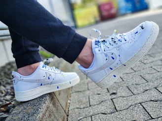 A pair of white sneakers with colorful paint splatters, worn by a person sitting on a low concrete ledge. The sneakers have a classic design with a textured sole, and the paint splatters include colors like blue, yellow, and black. The person is wearing dark pants, and the background features a paved area and some blurred greenery.
