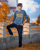 A young person is leaning casually against a concrete railing, wearing a blue checked long-sleeve shirt with yellow lettering and dark jeans. They are also wearing sunglasses and sneakers, with their hair styled neatly. In the background, there are vividly colored orange-brown trees and a building with a pink roof under a partly cloudy sky.