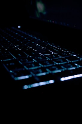 A close-up view of a computer keyboard, partially illuminated in a dark environment, highlighting the keys with a subtle glow.