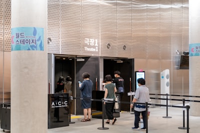 People are entering a theater or event space with signs in Korean and English. The area appears organized with ropes guiding attendees. A staff member is assisting, and the environment looks modern with metallic and glass elements.