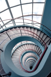 A spiral staircase viewed from above, showcasing its circular, symmetrical design with metal railings and steps. The staircase is enclosed by a large window structure, allowing natural light to flood the space. The railings are painted in a combination of red and blue colors.