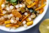 A close-up of a bowl filled with a mixture of white sago pearls, golden raisins, green chili slices, and yellow fried potato sticks. A curry leaf is visible, adding a hint of green. A lemon slice is partially visible next to the bowl.