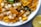 A close-up of a bowl filled with a mixture of white sago pearls, golden raisins, green chili slices, and yellow fried potato sticks. A curry leaf is visible, adding a hint of green. A lemon slice is partially visible next to the bowl.