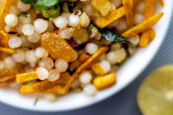 A close-up of a bowl filled with a mixture of white sago pearls, golden raisins, green chili slices, and yellow fried potato sticks. A curry leaf is visible, adding a hint of green. A lemon slice is partially visible next to the bowl.