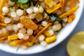 A close-up of a bowl filled with a mixture of white sago pearls, golden raisins, green chili slices, and yellow fried potato sticks. A curry leaf is visible, adding a hint of green. A lemon slice is partially visible next to the bowl.