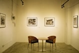 A minimalist art gallery setting features a small round table with two curved wooden chairs. The walls display several black and white framed photographs. The room is illuminated by track lighting, creating a serene and contemplative atmosphere.
