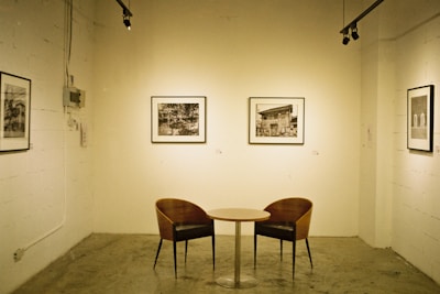 A minimalist art gallery setting features a small round table with two curved wooden chairs. The walls display several black and white framed photographs. The room is illuminated by track lighting, creating a serene and contemplative atmosphere.
